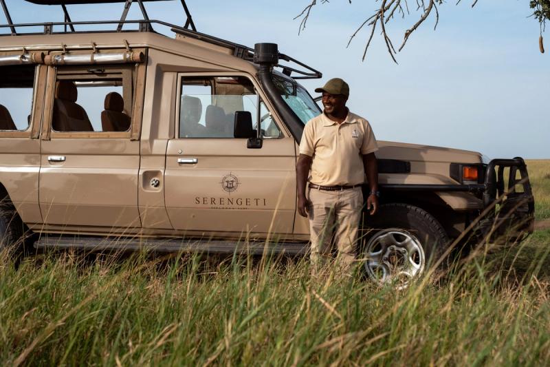A Serengeti Sojourn Camp guide standing beside a safari vehicle in the grassy plains of Tanzania’s Serengeti.