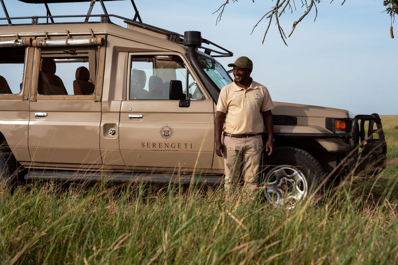 A Serengeti Sojourn Camp guide standing beside a safari vehicle in the grassy plains of Tanzania’s Serengeti.