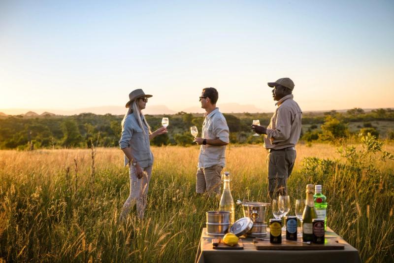 A couple on their African safari honeymoon savouring sundowners in a golden grassland setting, surrounded by the serene beauty of the African bush at sunset