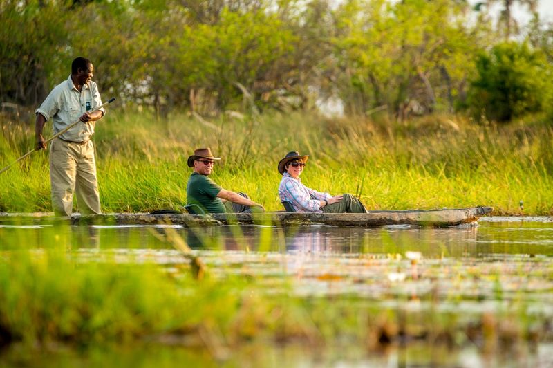 Two travellers glide in a mokoro canoe through calm, reed-fringed waters, experiencing the tranquillity of Africa in September.