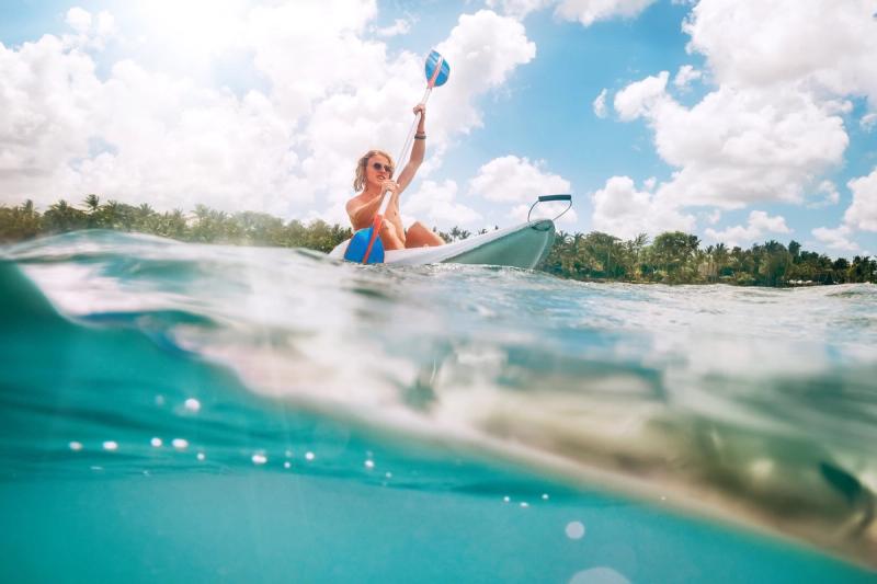 A teenager paddles a kayak in turquoise waters near a palm-fringed shore, capturing how beach destinations for kids can still feel cool enough for teens.