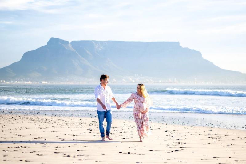 Katharina in a flowing floral dress walks hand-in-hand with her husband along a sandy beach, with Table Mountain rising in the background during their babymoon safari.