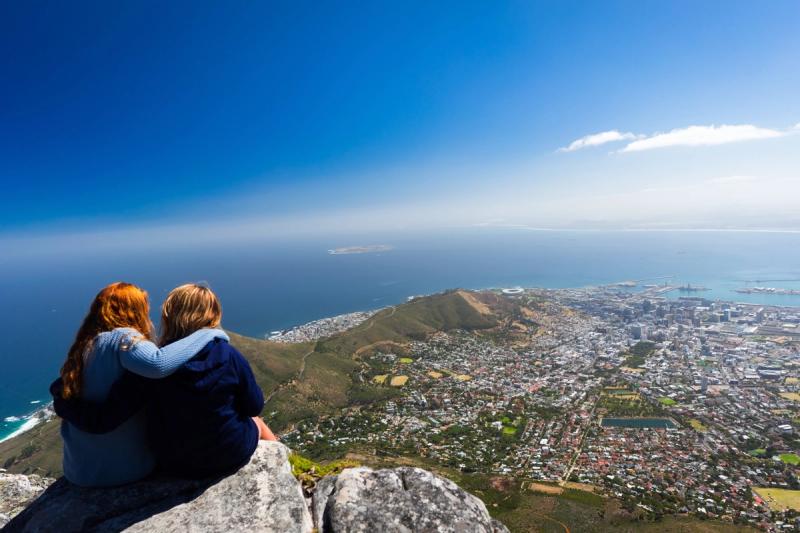 Two young adults sit on a mountaintop overlooking Cape Town and the ocean, reflecting how destinations for kids can grow into places of independence and perspective.