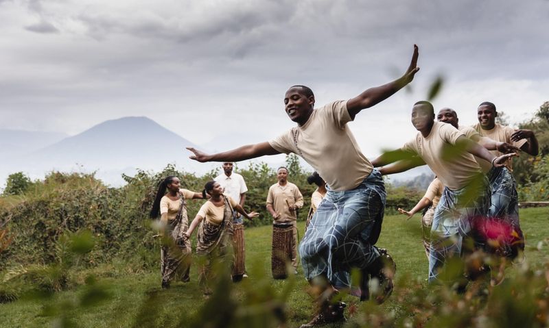 A group of Rwandan dancers perform joyfully outdoors with mountains in the distance, their movements full of energy and pride.