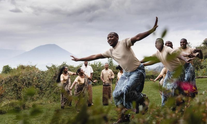 A group of Rwandan dancers perform joyfully outdoors with mountains in the distance, their movements full of energy and pride.