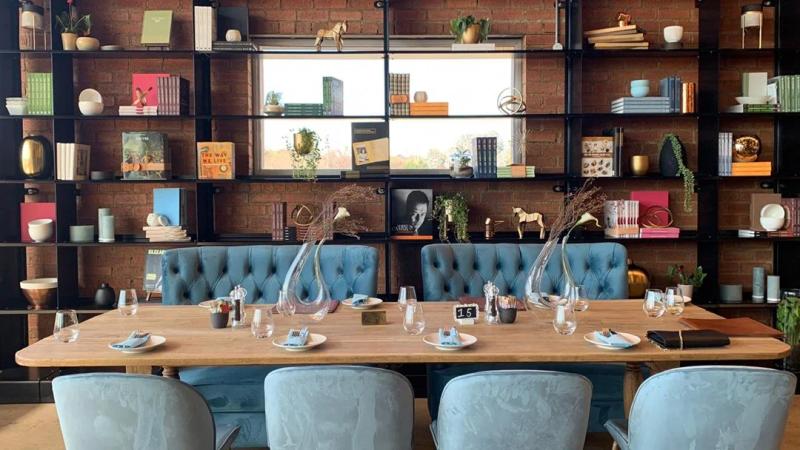 A long dining table with place settings and blue couches and chairs around it and a bookcase behind it