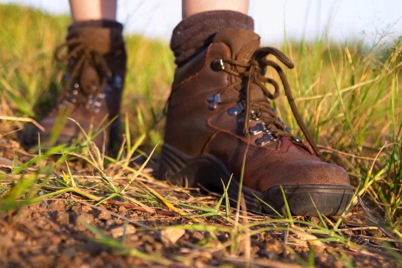 A close-up of sturdy brown hiking boots stepping on dry grass and dirt in the sunlight.