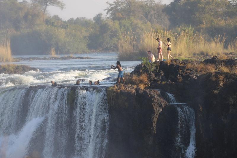 A group of swimmers relaxes in Devil’s Pool while others watch from the rocky edge above the best side of Victoria Falls.