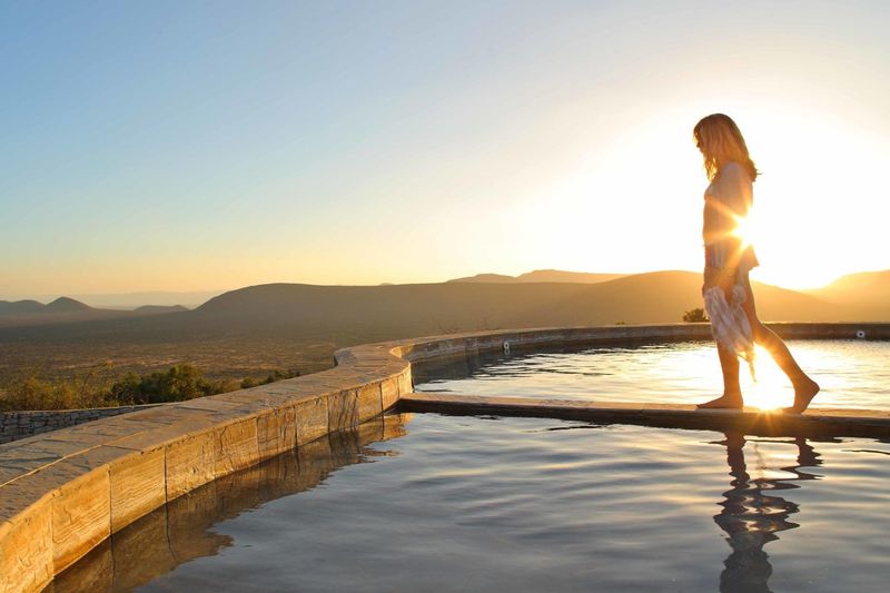 A woman walks along the edge of a round pool at sunset, overlooking rolling hills – a quiet moment that still counts as the best time to visit Kenya.