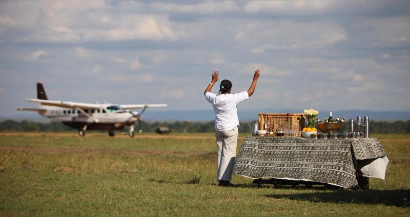 A camp staff member waves to arriving guests stepping off a small bush plane beside a table laid out with refreshments on a grassy airstrip.