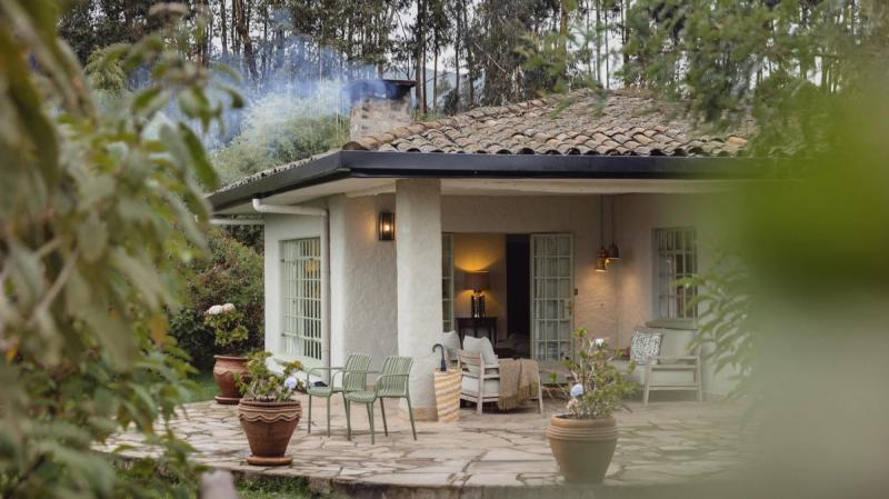 A cosy cottage with soft lighting, outdoor seating, and smoke rising from the chimney is surrounded by potted plants and leafy trees.