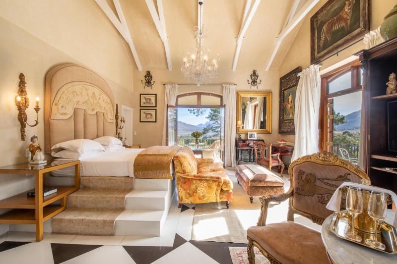 An elegant and opulent bedroom with a plush bed on raised steps, antique furniture, chandeliers, ornate décor, and large windows offering stunning views of the surrounding mountains