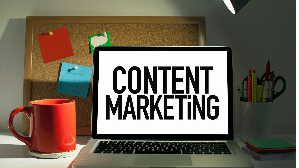 "A laptop on a desk displaying the words 'Content Marketing,' with a red mug, stationery, and a corkboard in the background, representing a workspace focused on creating content marketing strategies.