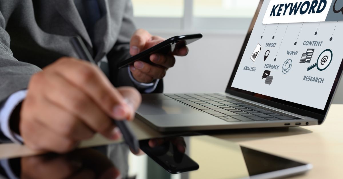 A person holding a smartphone while working on a laptop displaying a keyword research and content strategy diagram.