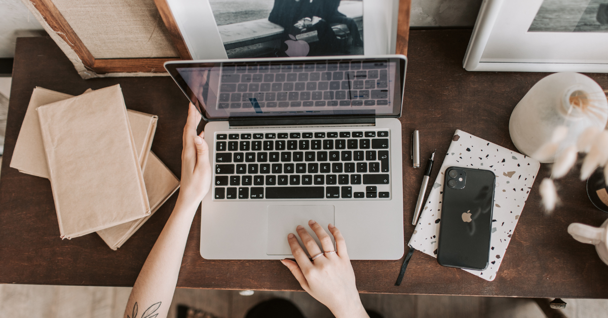 erson working on a silver MacBook laptop with an iPhone on a rustic wooden desk, surrounded by business notes and craft paper stacks.