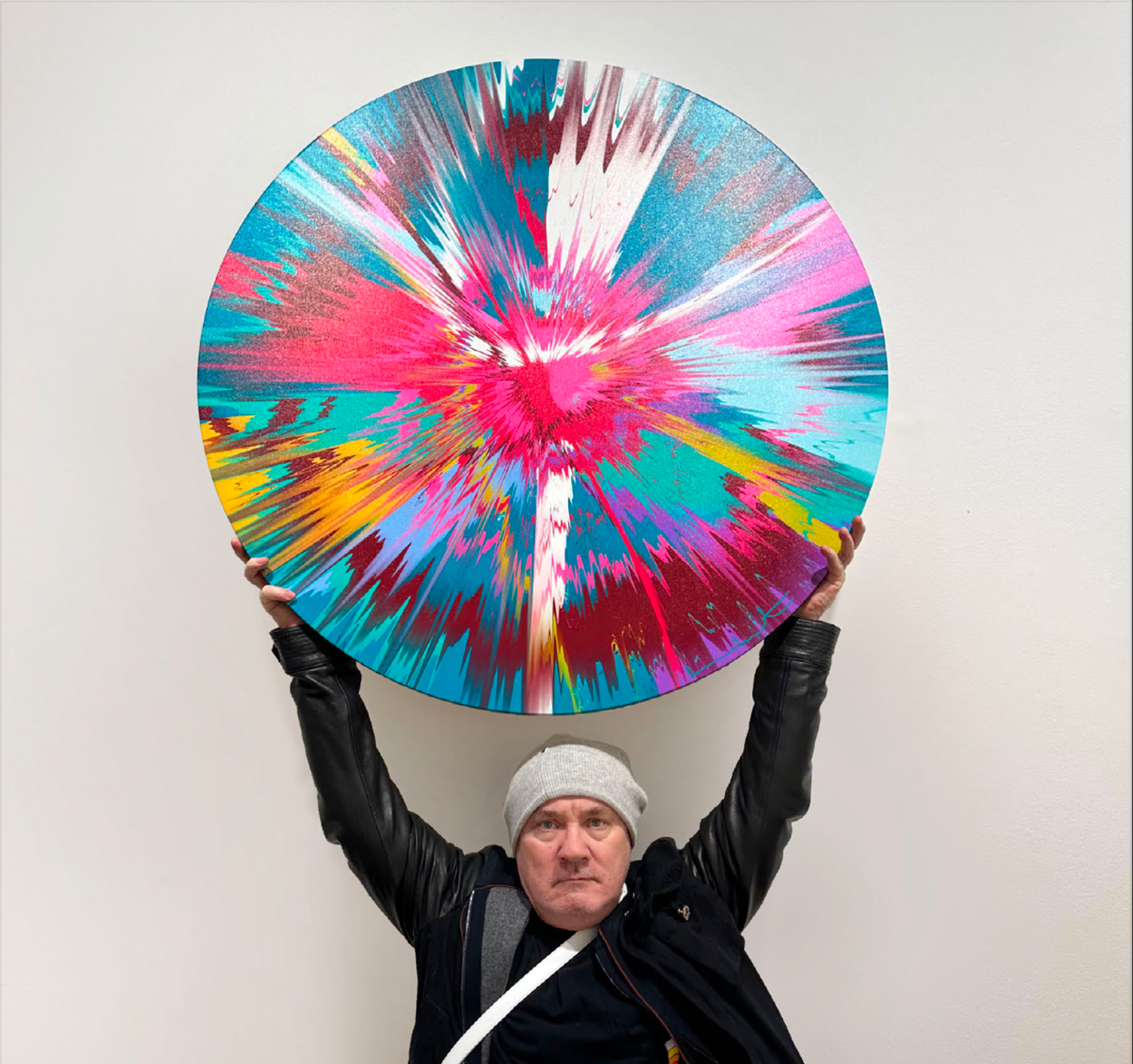 Artist Damien Hirst, dressed in a black leather jacket, holding one of his circular Spin Paintings above his head. The Spin Painting is composed of a varied palette of blues, pinks, reds, yellows, and whites, in a dizzying pattern.