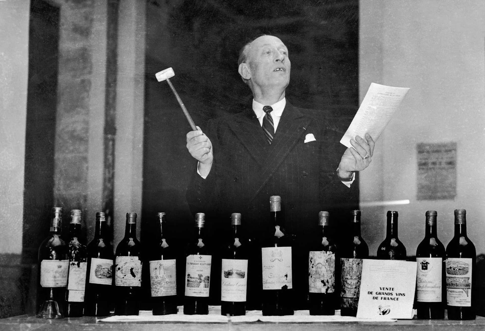 Black and white image of a male auctioneer speaking out to the room with a row of wines in front of him holding a gavel in one hand and a paper sheet in the other