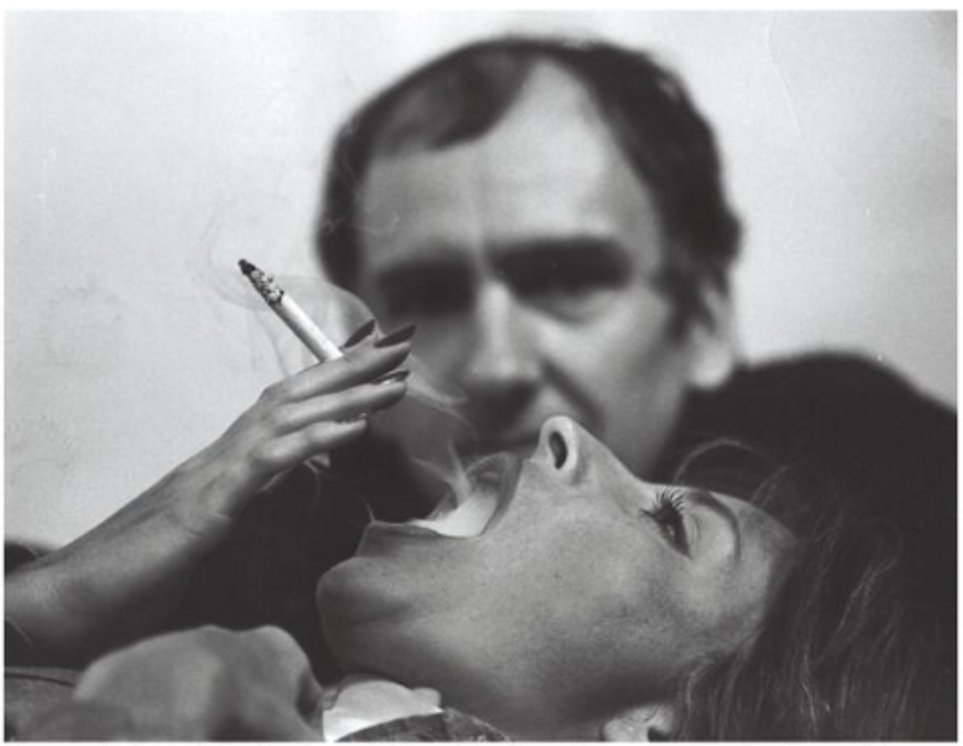 Photograph of Tom Wesselmann holding a cigarette above a reclining woman's mouth, capturing the moment of smoke exhalation.