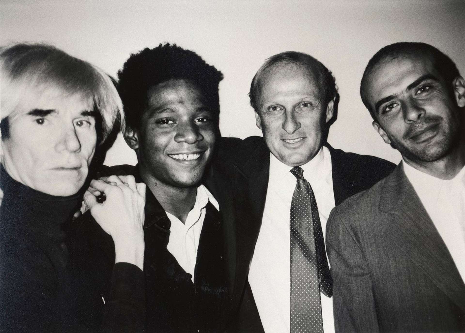 A black and white photograph of  Andy Warhol, Jean-Michel Basquiat, Bruno Bischofberger and Fransesco Clemente, New York, 1984.