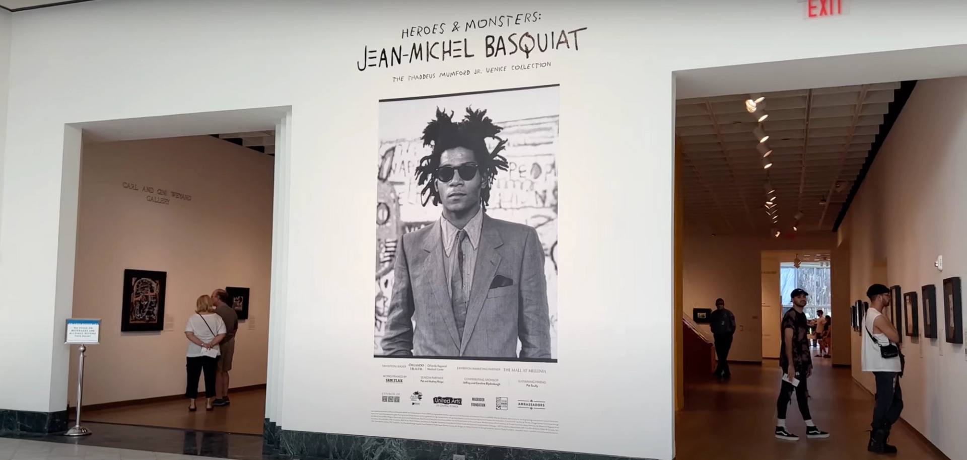 A screenshot of a Youtube video showing the installation view of Basquiat's Heroes and Monsters exhibit. It shows a larger-than-life black and white portrait of Basquiat, wearing a suit and sunglasses, against a white wall. On either side of the wall, visitors to the exhibition look at the artworks.