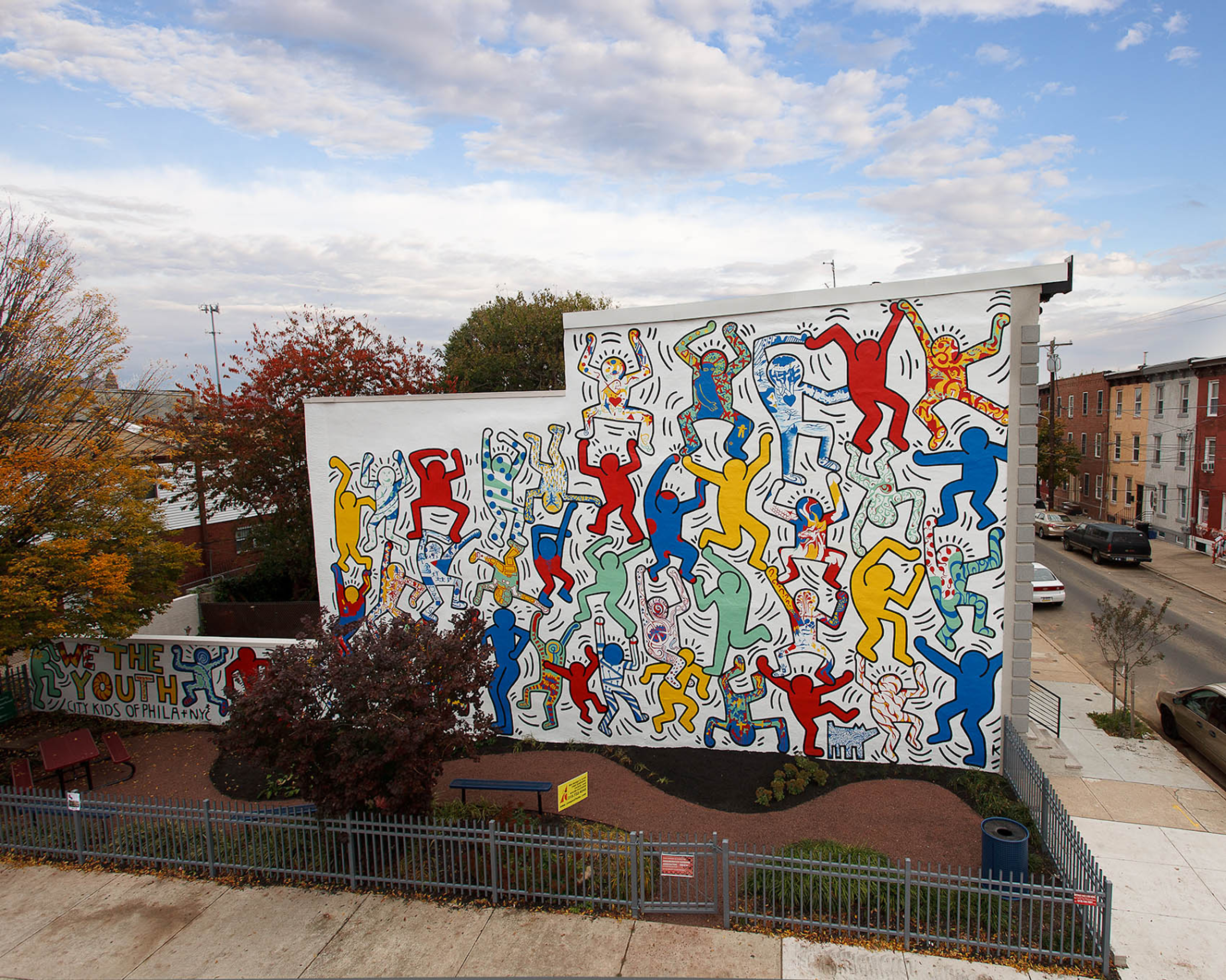  Photograph of Keith Haring's We The Youth mural. A white mural with a dog and red, blue, yellow and green figures displaying a variety of movements. 