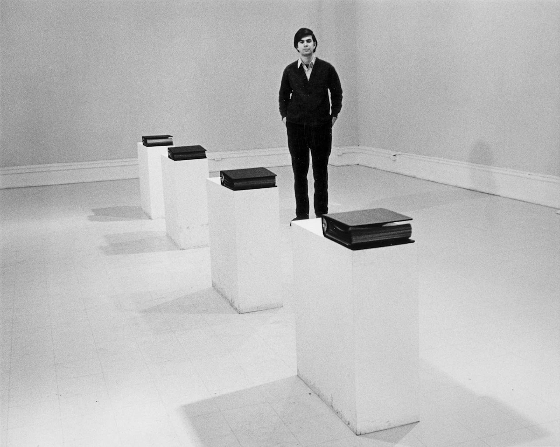 A black and white photographic image of mel bochner standing amoung four plints each holding a 