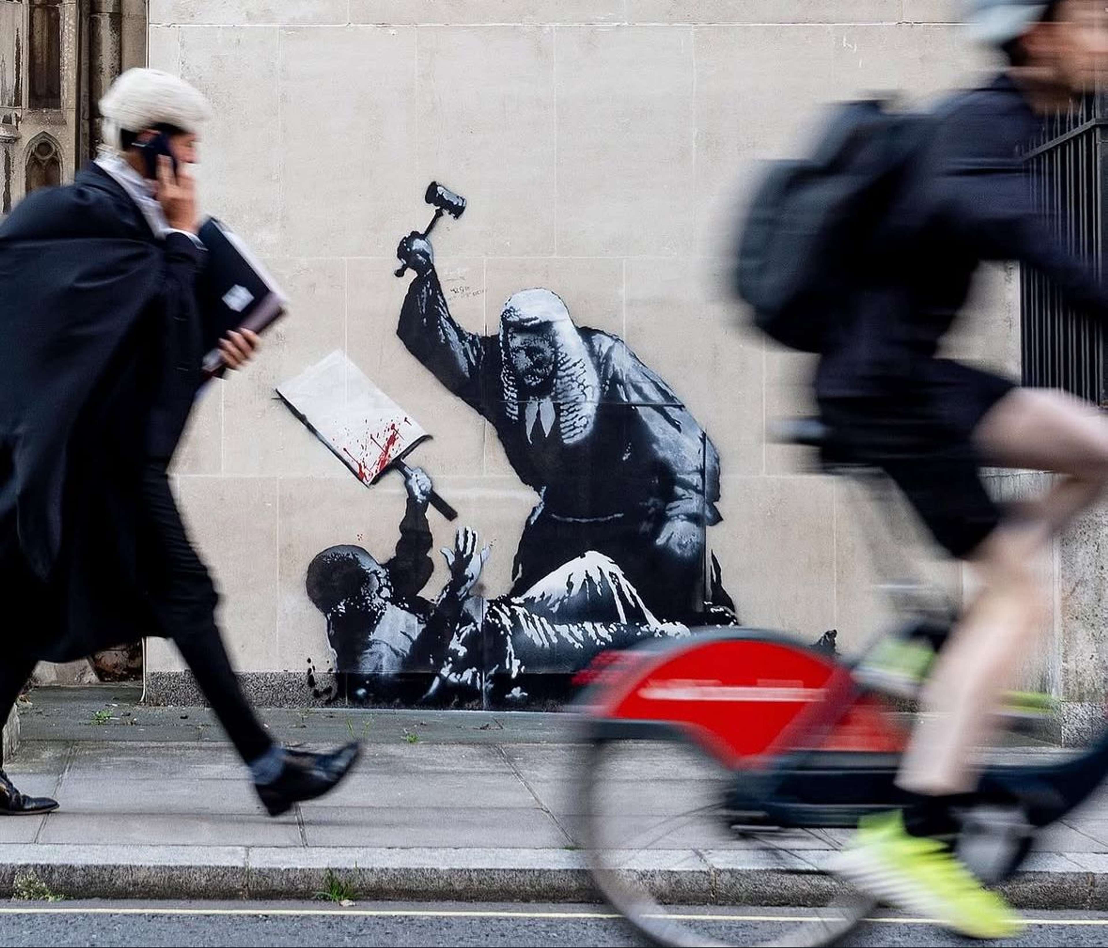 A black-and-white stencil mural on a pale stone wall shows a robed judge raising a gavel over a protester lying on the ground, as blurred cyclists and pedestrians rush past in the foreground.