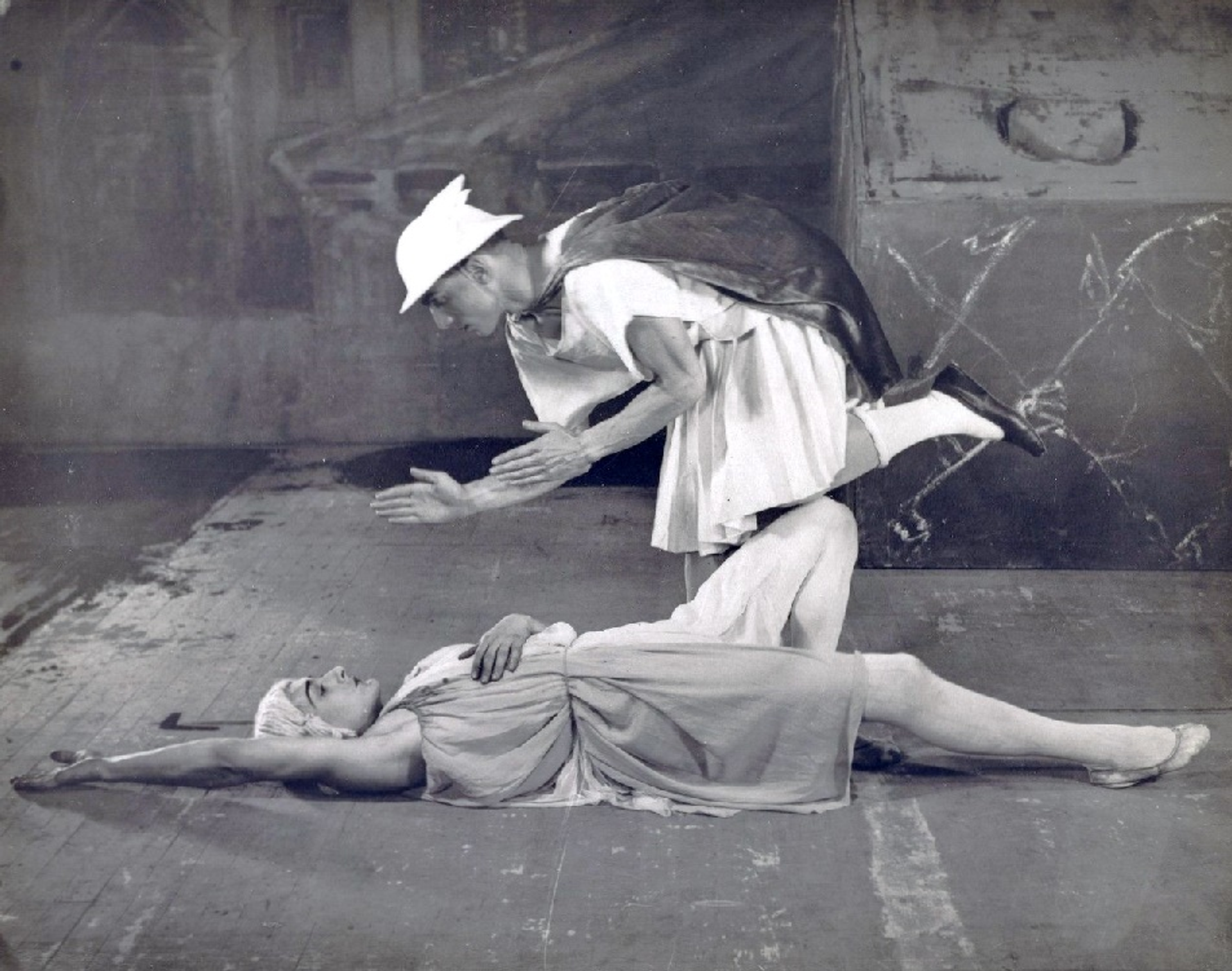 A monochrome image showing Massine being killed by Mercury, shown wearing a tunic and his signature hat. She is shown lying on the floor with her arms extended over her head.