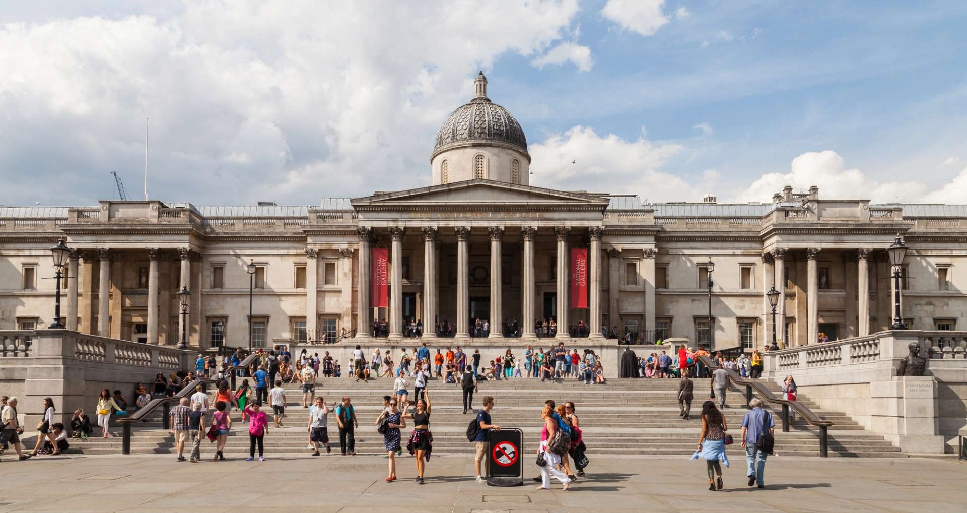 An image of the National Gallery, London from the outside. It shows the iconic building, seen from Trafalgar Square on a bright and sunny day. Many people stand on the steps.