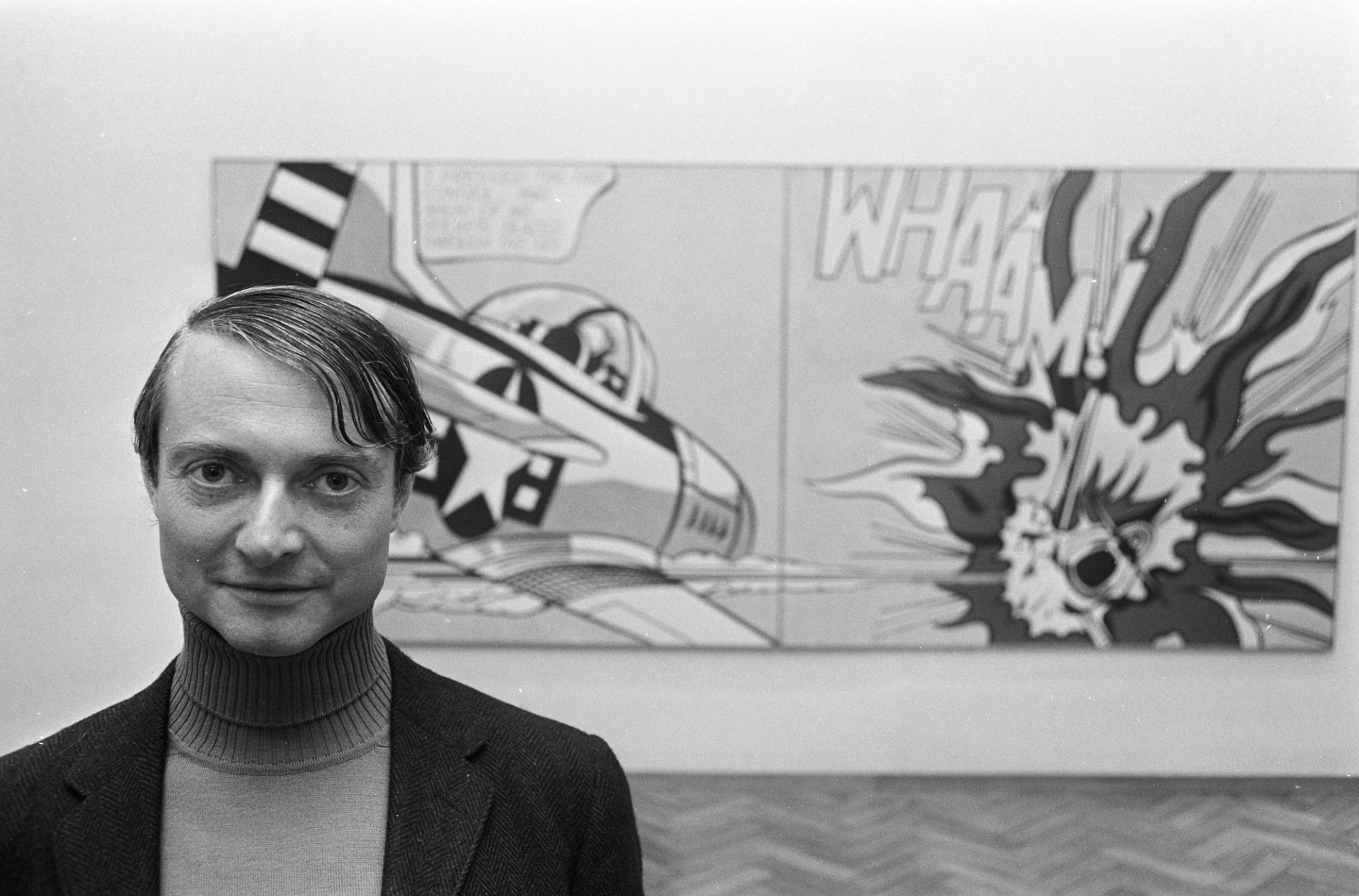 A monochrome portrait of Roy Lichtenstein, wearing a suit and standing in front of his iconic work Whaam!