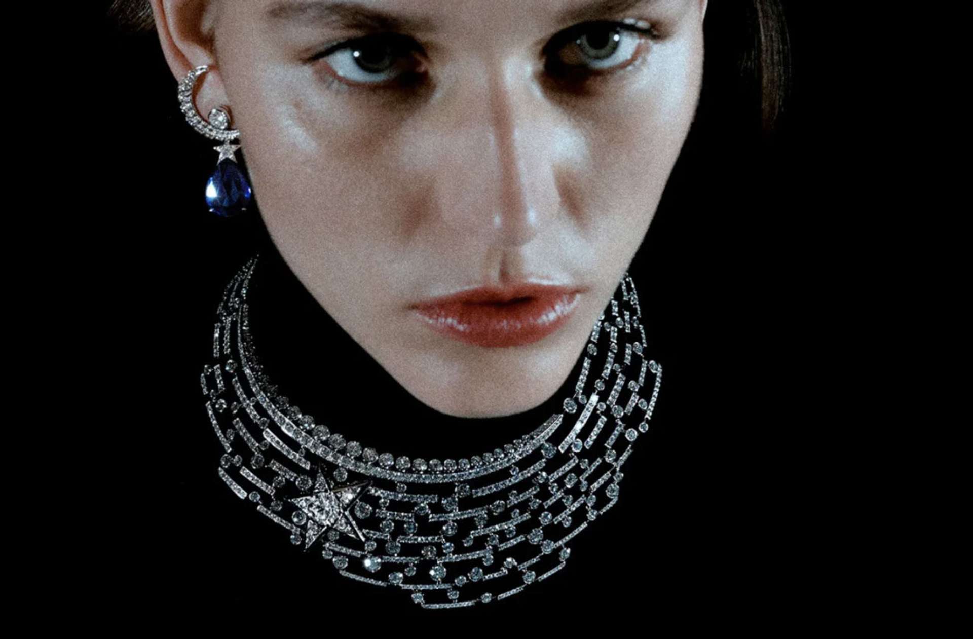 Woman wearing a thick silver choker necklace made up of links with a bejewelled star on one side, and earrings in the shape of a moon and a blue stone dropping from a connected star