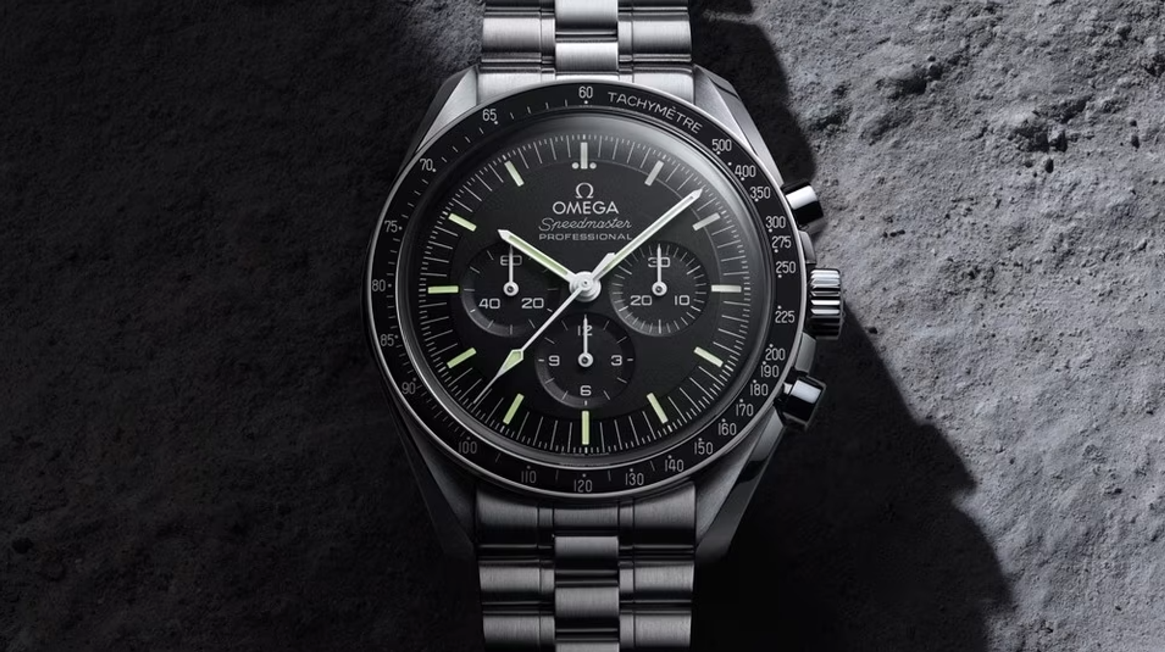 A black Omega Speedmaster, 1957. Set against a rocky background, the watch strap and setting is silver metal with a black face and three subdials.