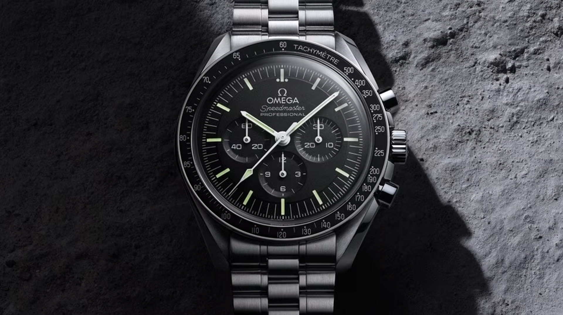 A black Omega Speedmaster, 1957. Set against a rocky background, the watch strap and setting is silver metal with a black face and three subdials.