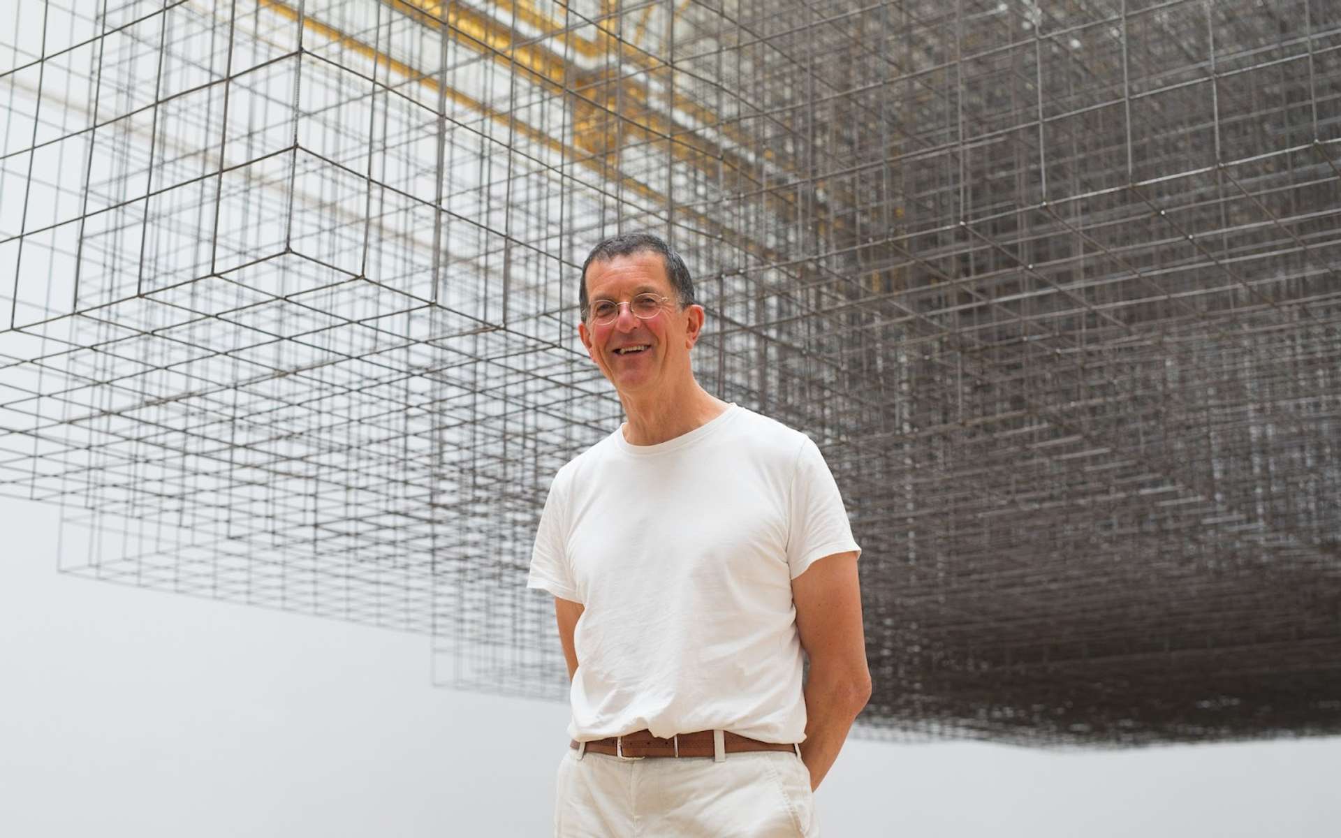  A picture of artist Antony Gormley standing in front of one of his works. He is wearing a white t-shirt and smiling at the camera.