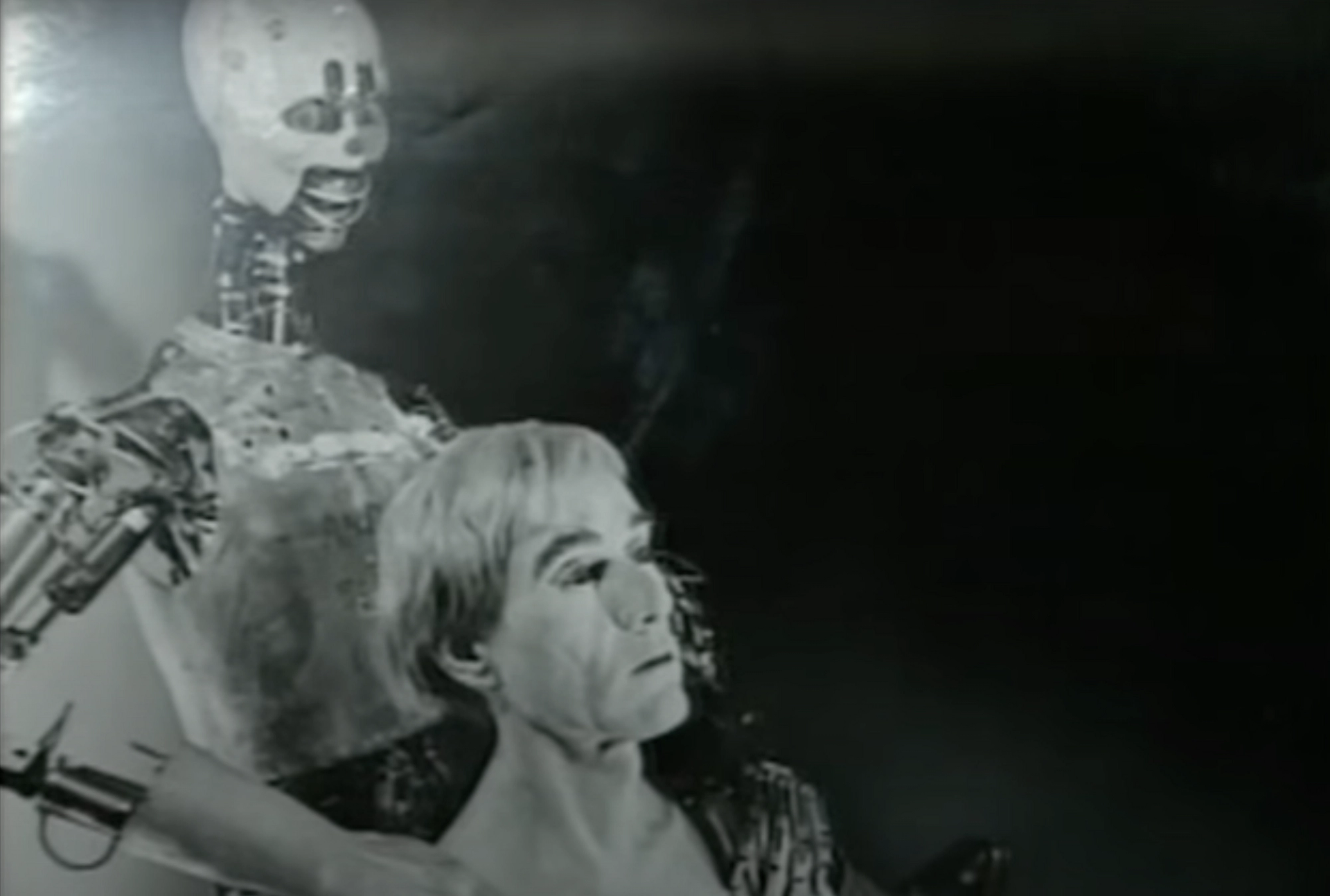 A screenshot of a video showing the face of Andy Warhol's 1981 Robot, in the likeness of the artist, being held by a larger metallic robot.