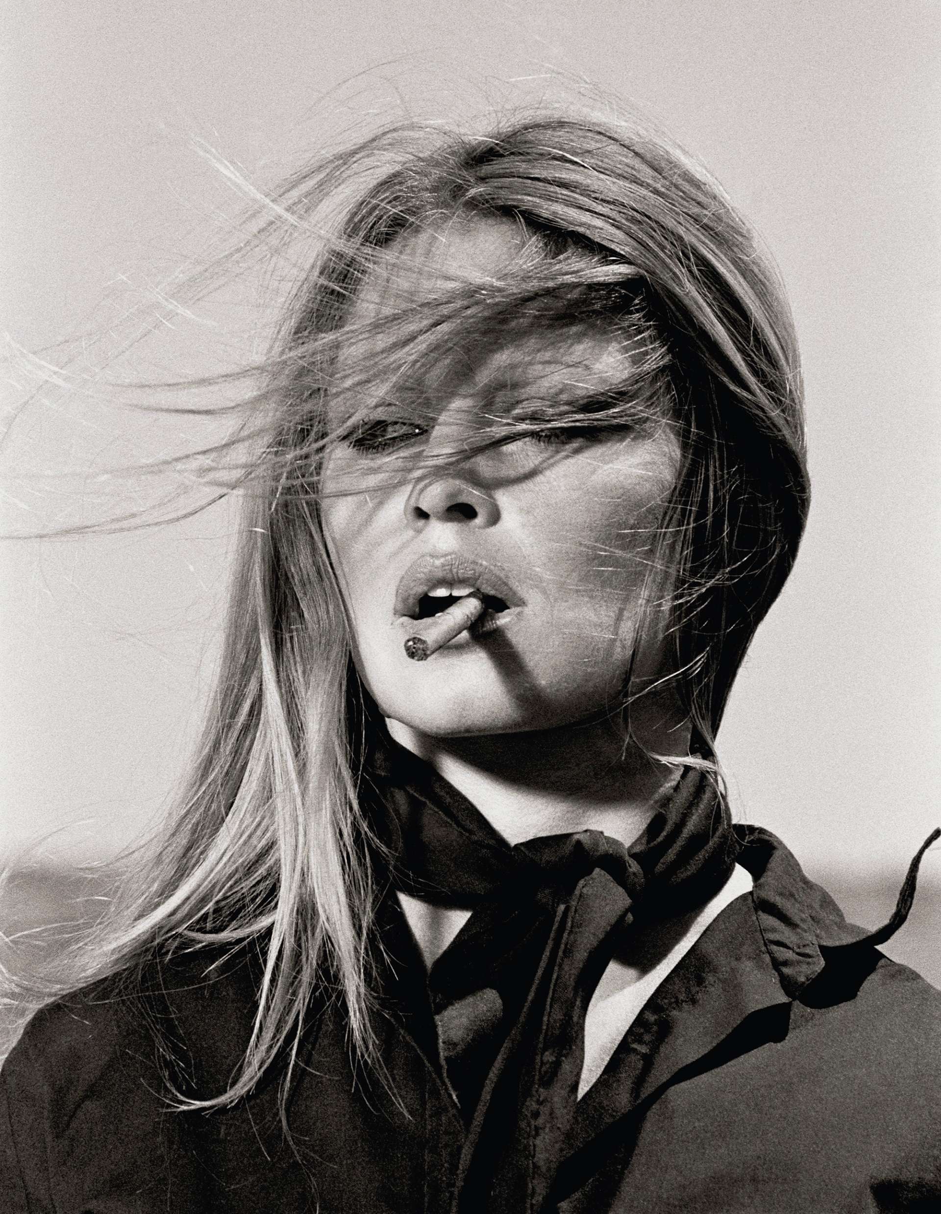 A black and white photograph by Terry O'Neill depicting Bridget Bardot's portrait at the centre of the shot, her hair windswept across her face. Bardot wears a black shirt and neckerchief, and has a lit cigarette between her lips.