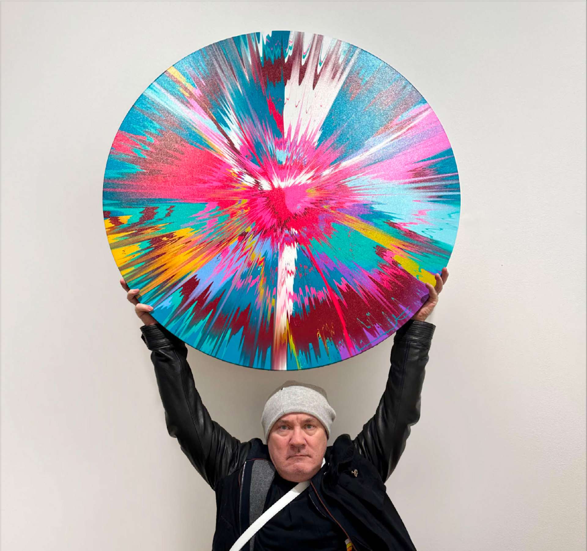 An image of the artist Damien Hirst holding an abstract, colourful round painting, part of his The Beautiful Paintings series.