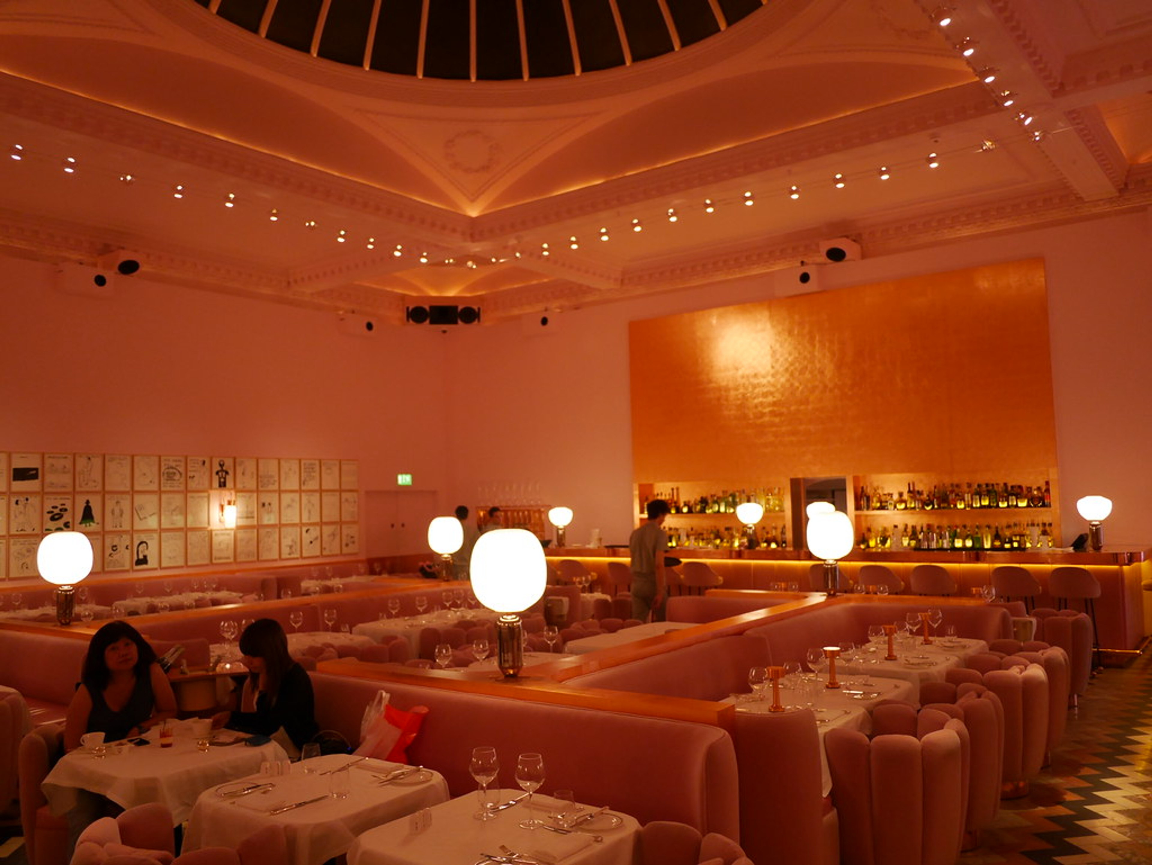 A view of the rose-gold hued restaurant Sketch, in London. Two women can be seen dining at a table on the bottom left corner.