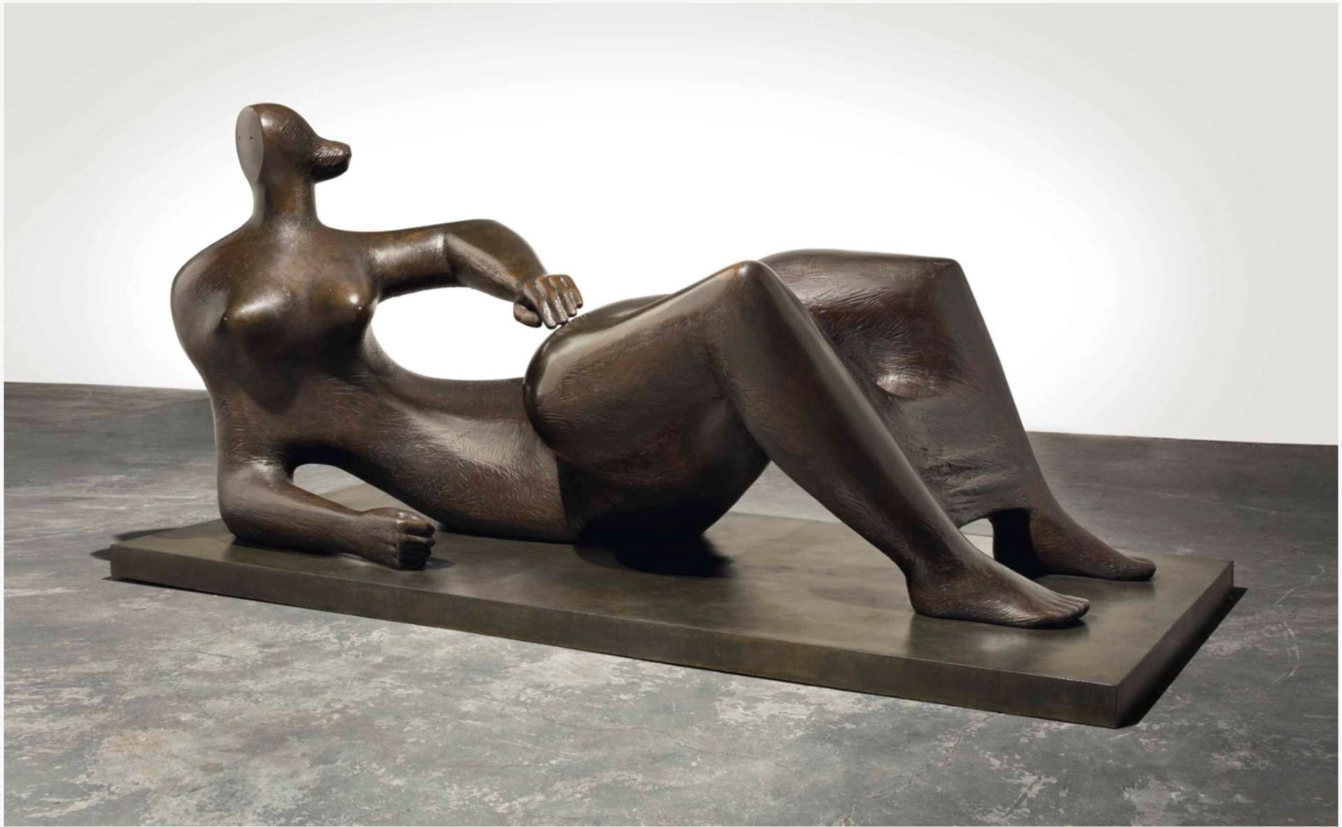  A large-scale bronze sculpture of a reclining biomorphic figure. The sculpture rests on a plinth, with the figure's feet firmly planted on the ground. One arm is raised in the air, while the elbow rests on the ground. The photograph captures the sculpture in an open white gallery space.