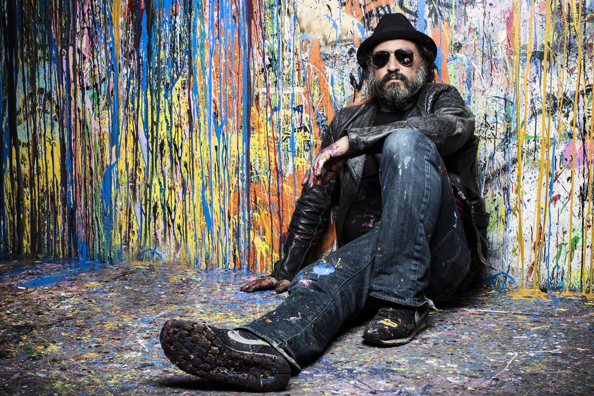A portrait of the artist Mr. Brainwash, showing wearing paint-splattered clothes and sunglasses. He is sitting on the floor and leaning against a wall full of colourful paint splatter.