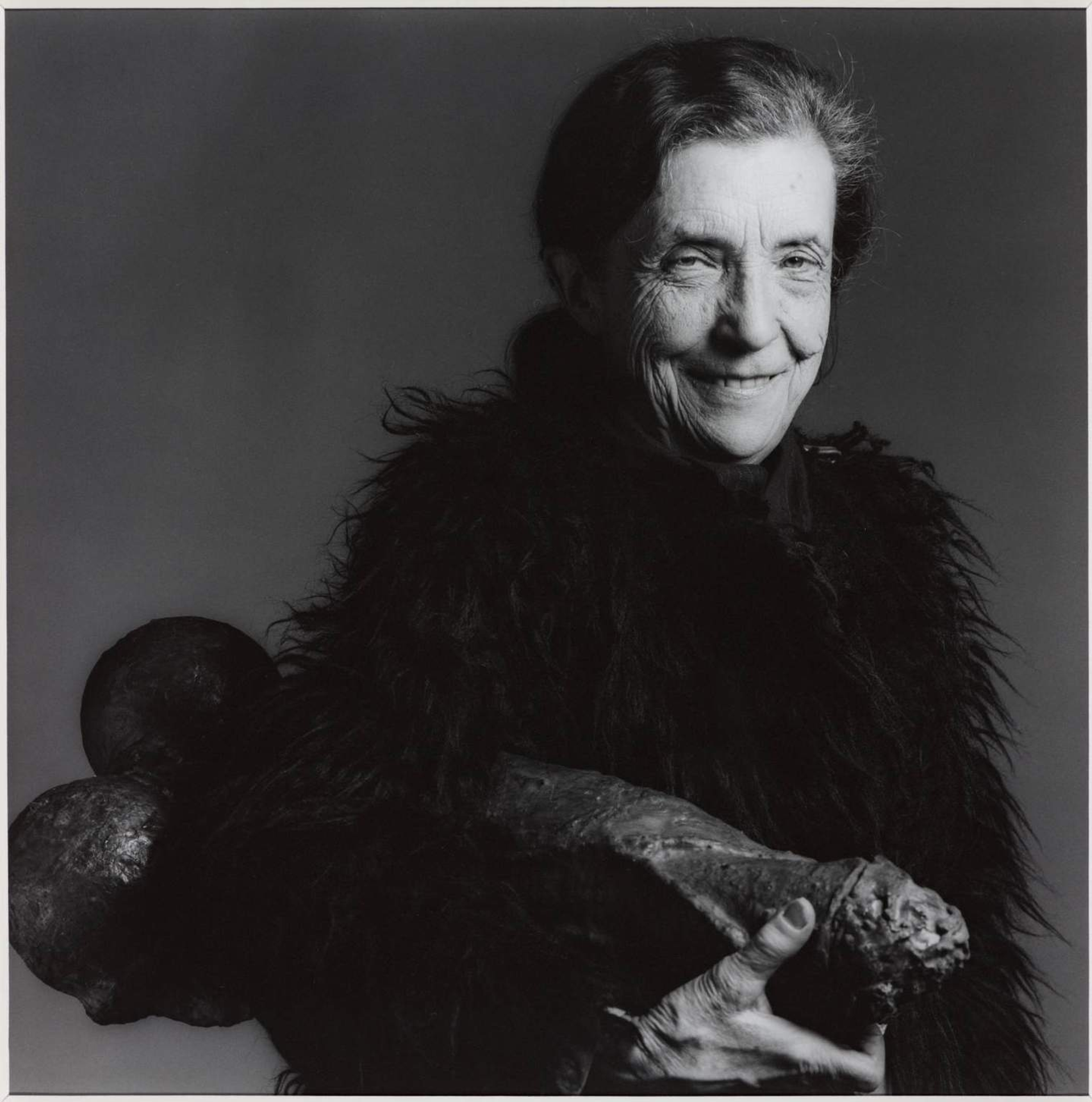 Black and white photograph of artist Louise Bourgeois smiling while holding onto a phallic shape object underneath her arm.