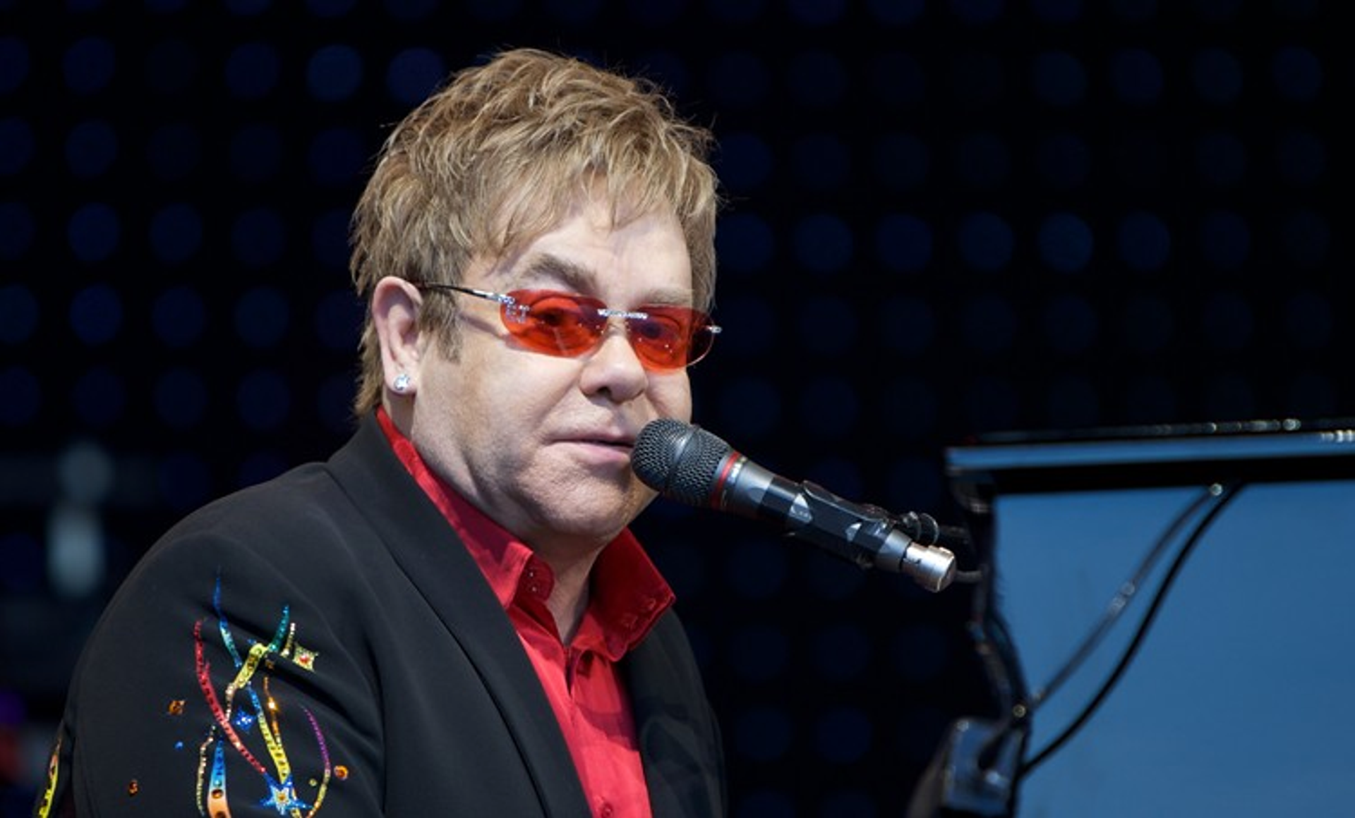 This photograph shows singer Elton John wearing his signature colourful glasses, sitting at a piano and singing into a microphone.