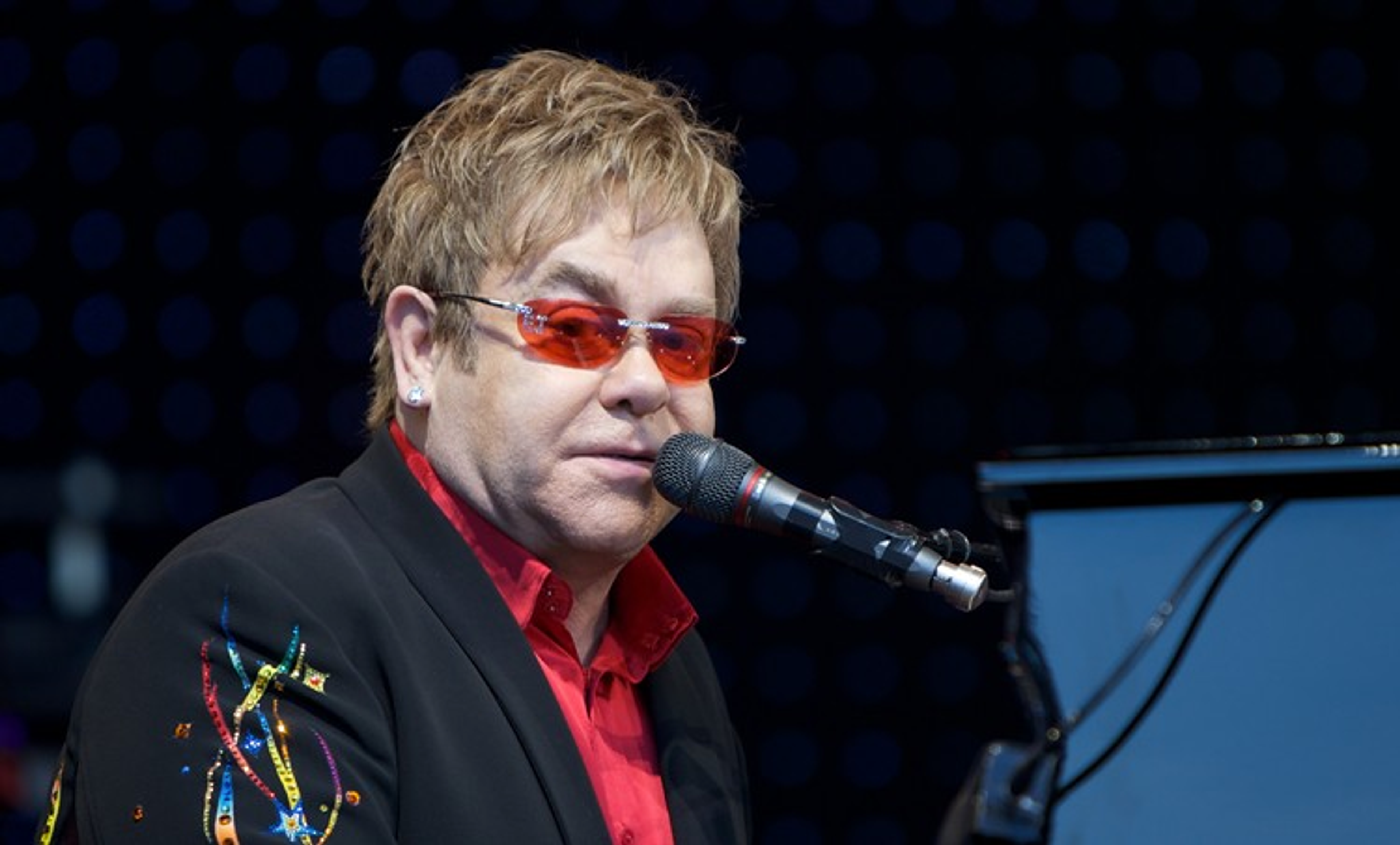 This photograph shows singer Elton John wearing his signature colourful glasses, sitting at a piano and singing into a microphone.