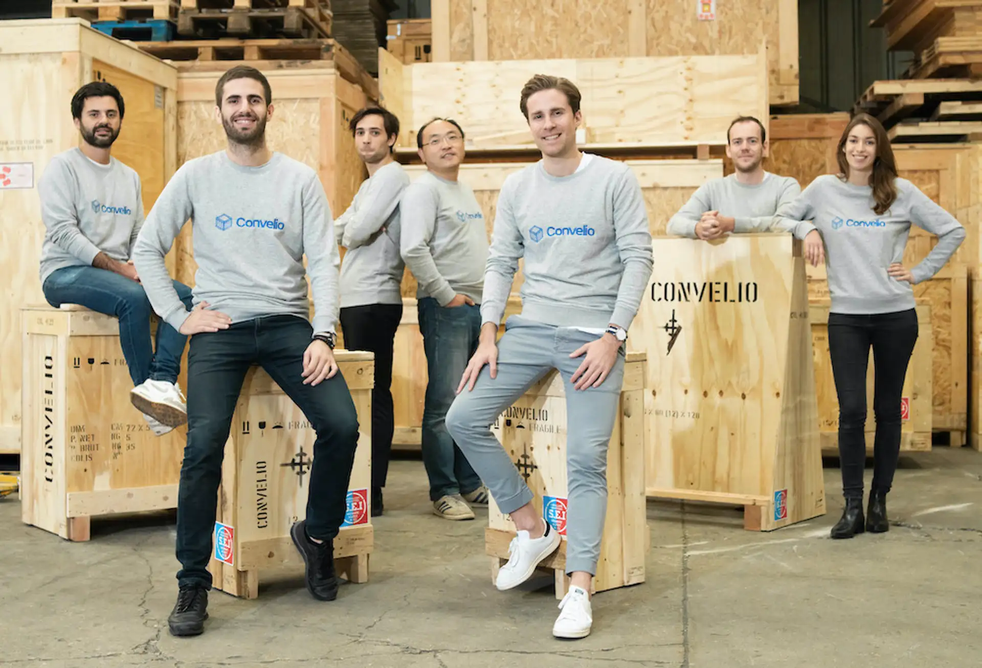 A photograph of the Convelio team, all wearing matching grey sweatshirts, leaning against wooden storage crates for art.