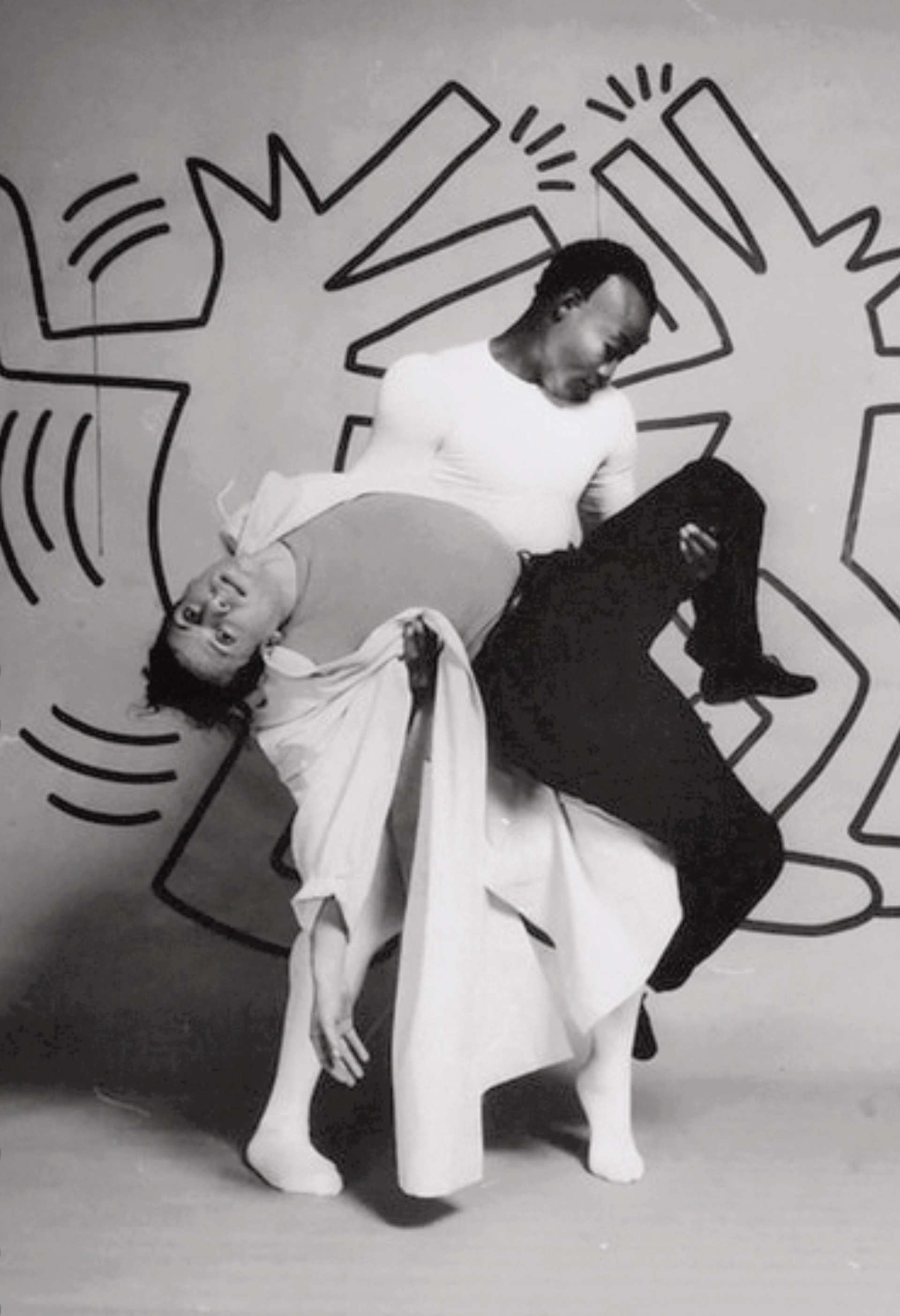Black and white photograph of the Secret Pastures set design. Two dancers posing in front of Keith Haring’s moving figures.