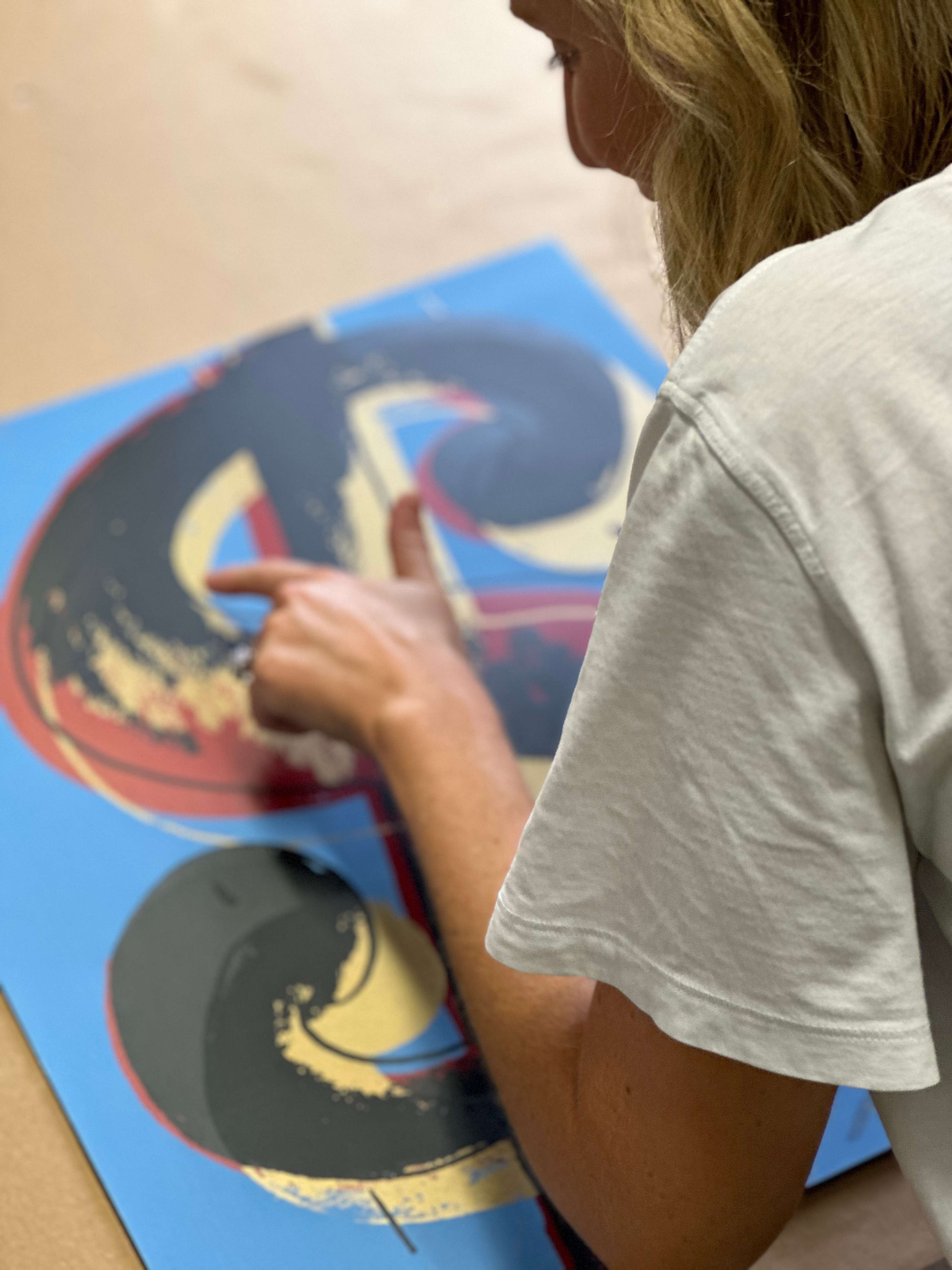 A blonde woman pointing at an artwork depicting a dollar sign in black, yellow, and red ink against a blue background