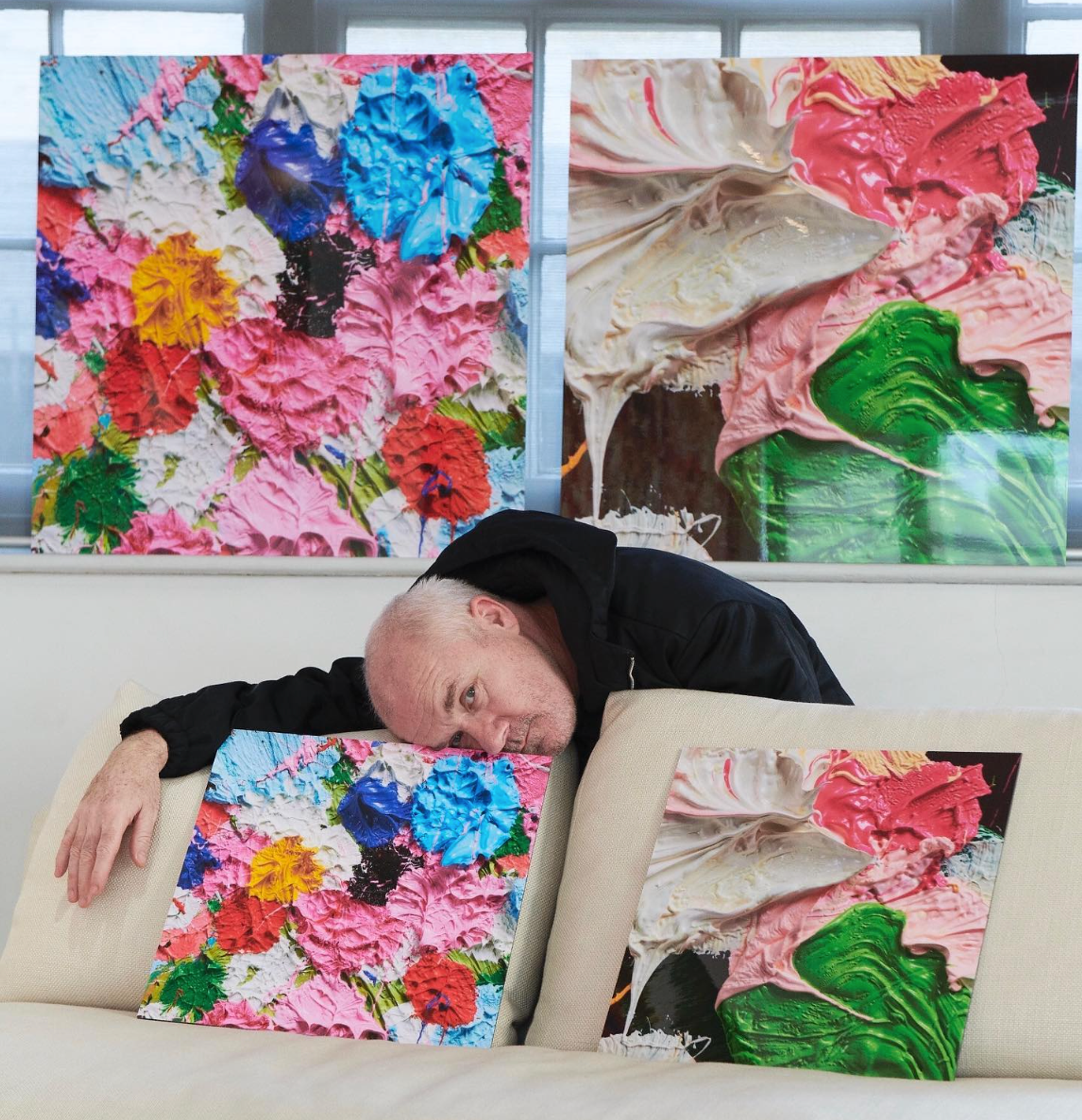 An image of the artist Damien Hirst, bent over a couch where two smaller versions of his works are perched. Behind him, two large-scale versions of Fruitful and Forever are visible.