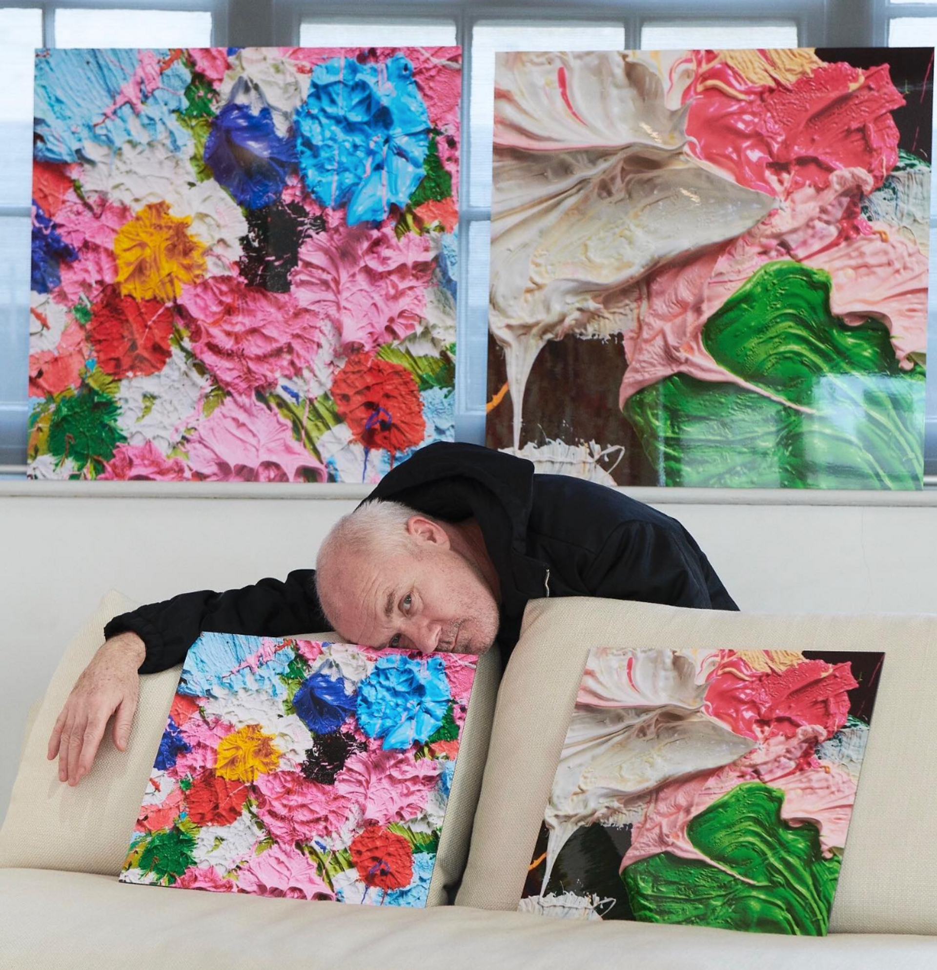 An image of the artist Damien Hirst, bent over a couch where two smaller versions of his works are perched. Behind him, two large-scale versions of Fruitful and Forever are visible.