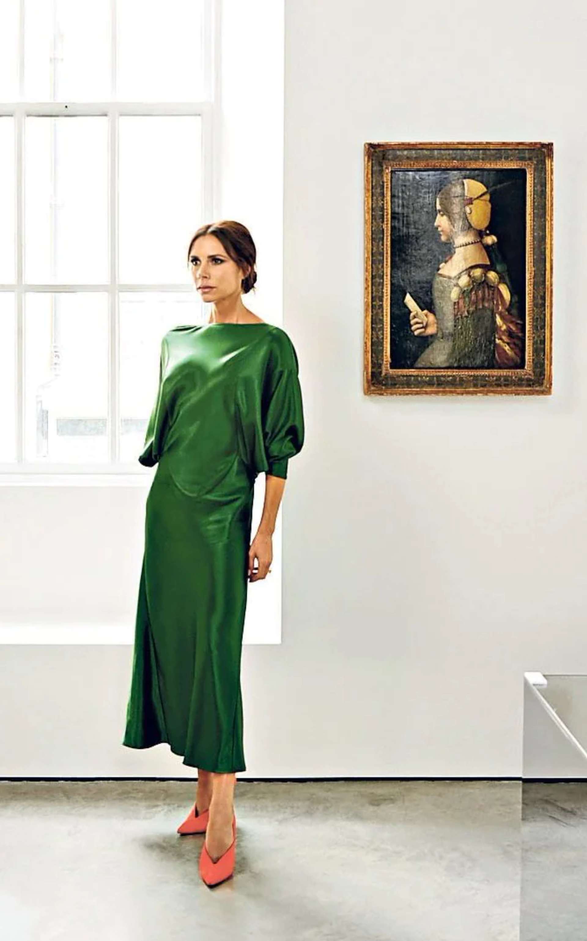 Victoria Beckham standing in her Dover Street store, wearing an emerald green silk dress and orange heels. Behind Beckham is a classic painting framed on the wall.