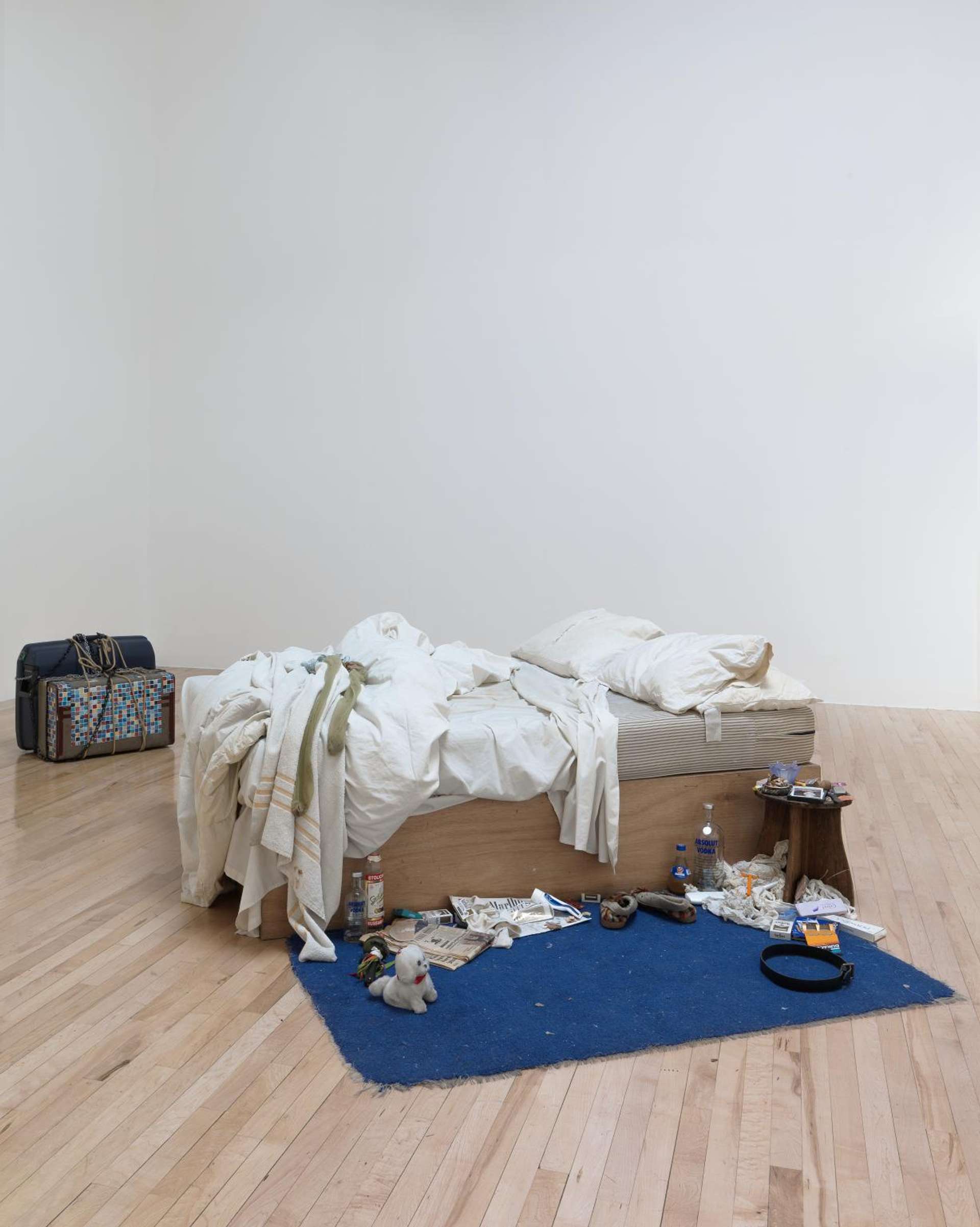 A photograph of My Bed (1998), an installation work by artist Tracey Emin. It shows a dishevelled bed, surrounded by empty alcohol bottles, cigarette butts and used condoms.